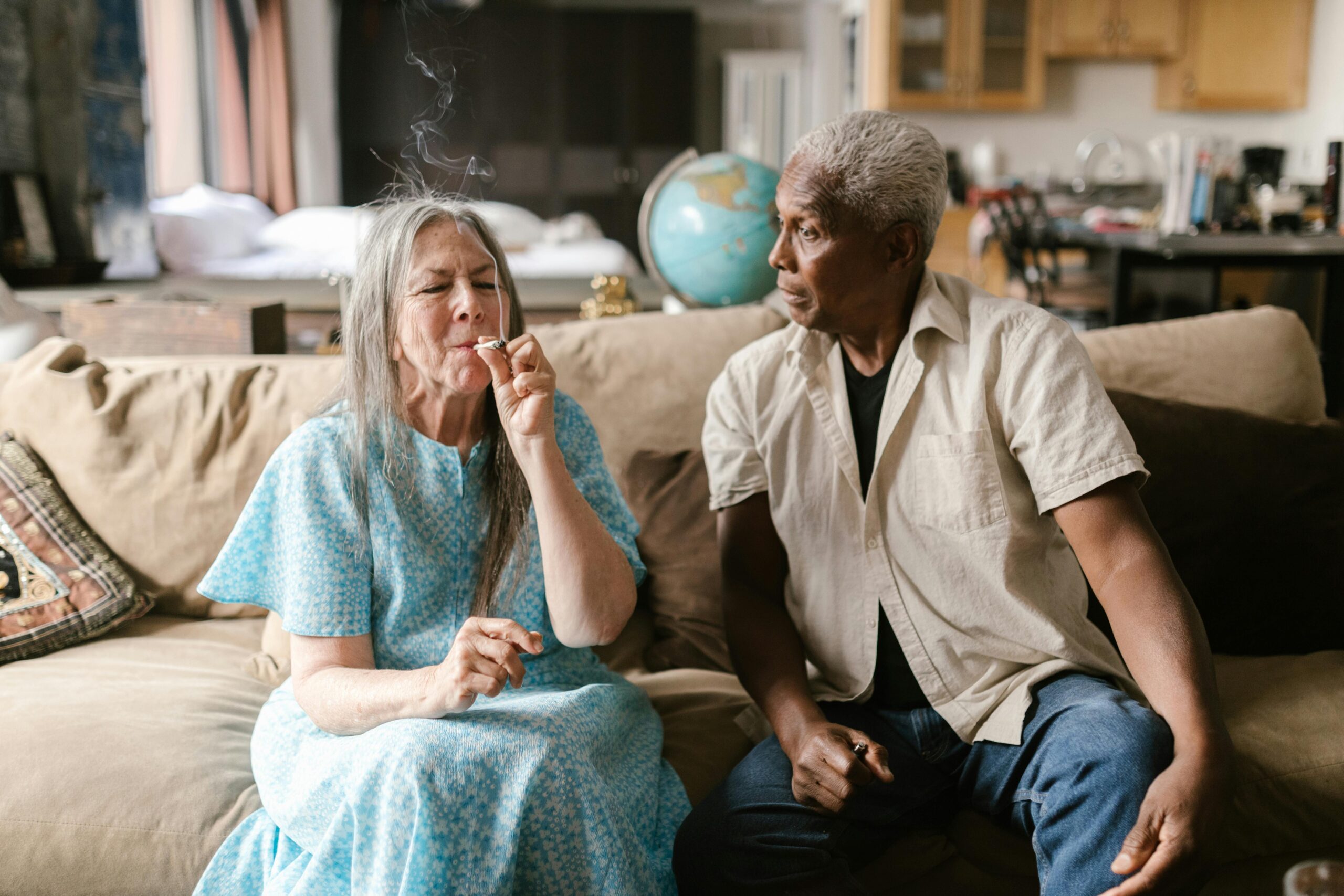 Interracial elderly couple sitting indoors, casually interacting with cannabis. Warm, relaxed setting.